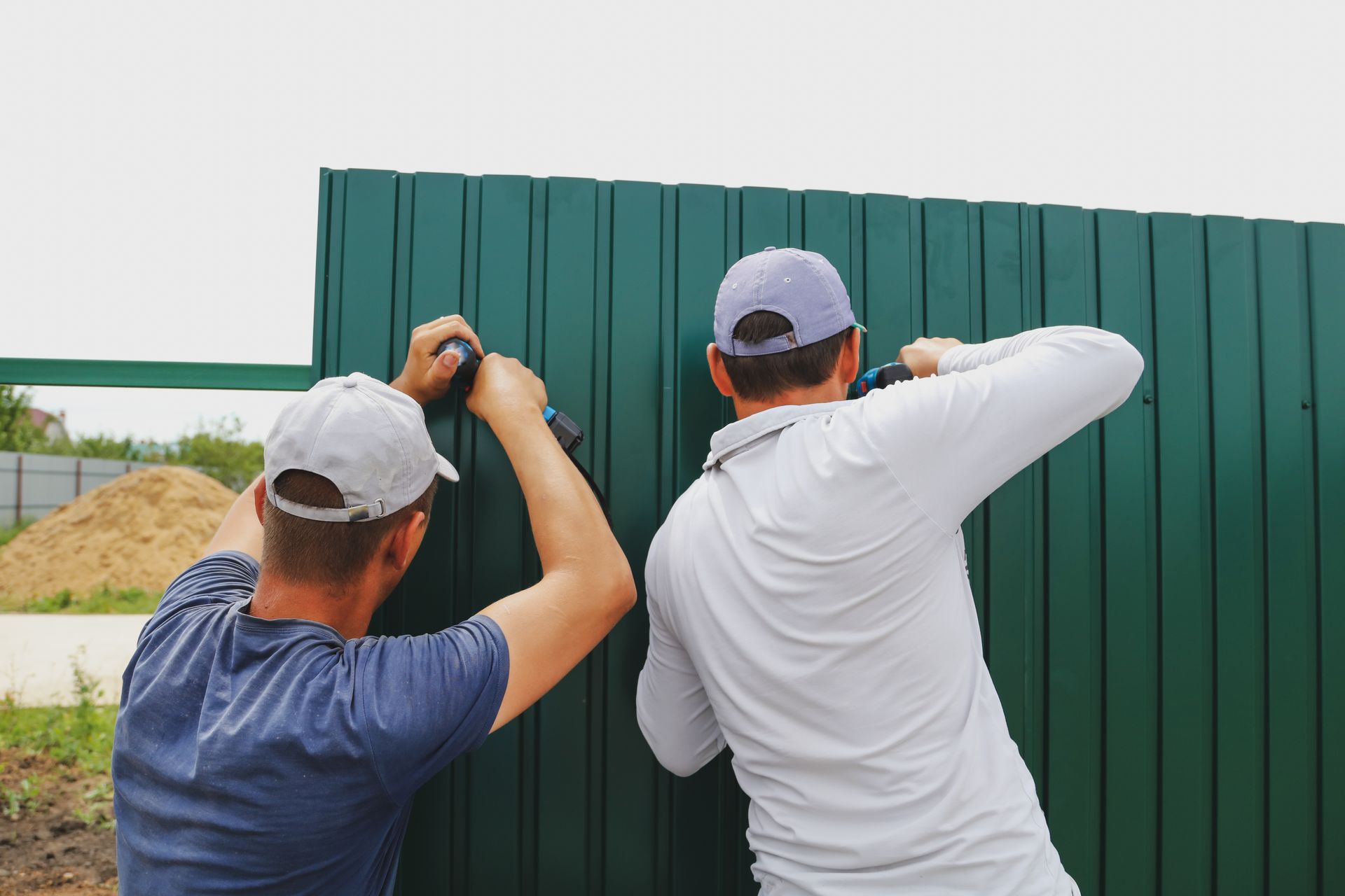 Two men in caps are installing a green metal fence using power tools outdoors.