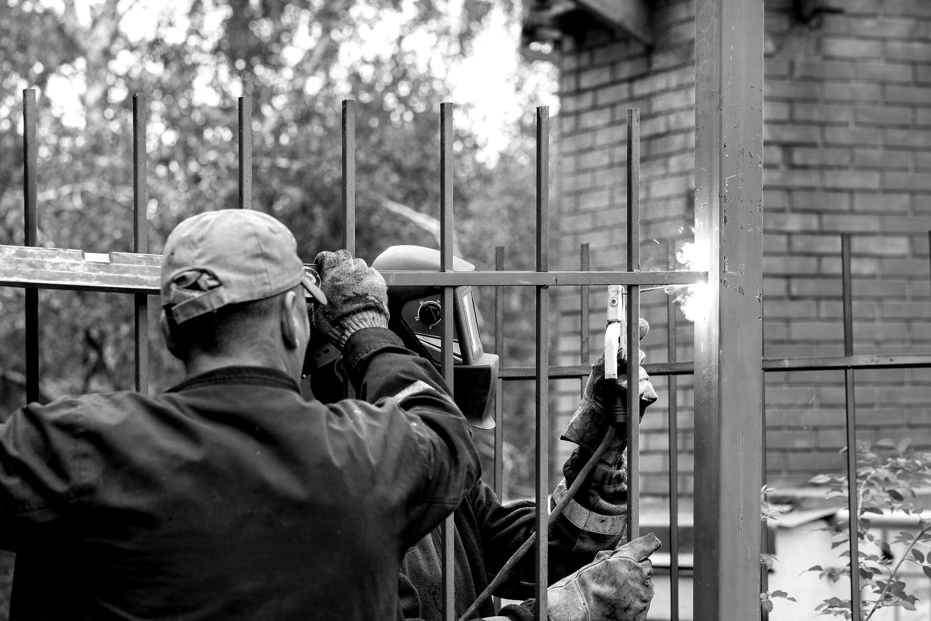 Two welders working on a black metal fence outdoors. One welding with sparks, the other assisting.