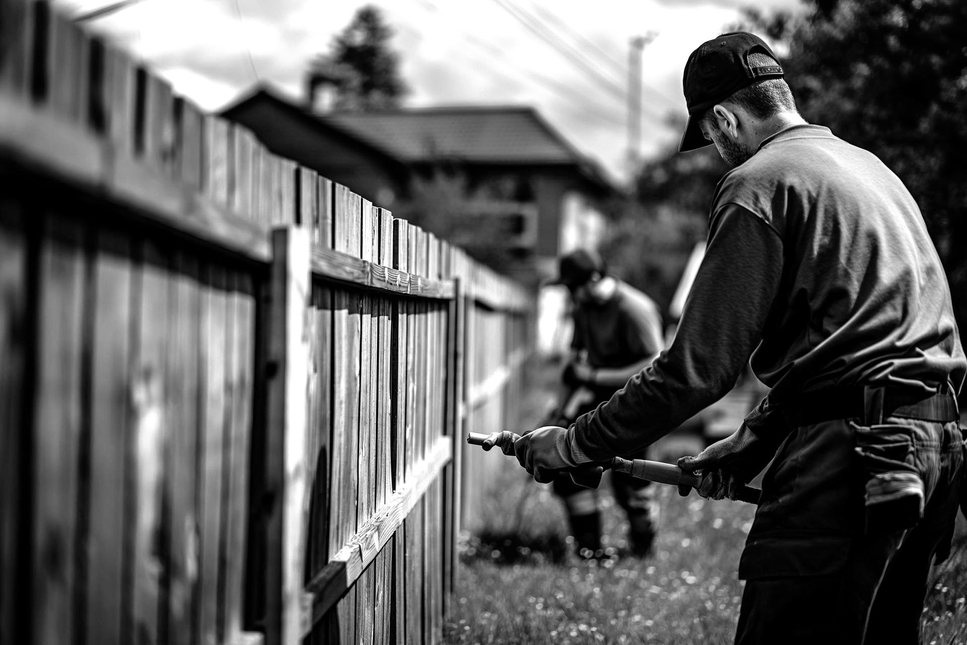 Two workers spraying a wooden fence with a paint sprayer outdoors. Black and white.