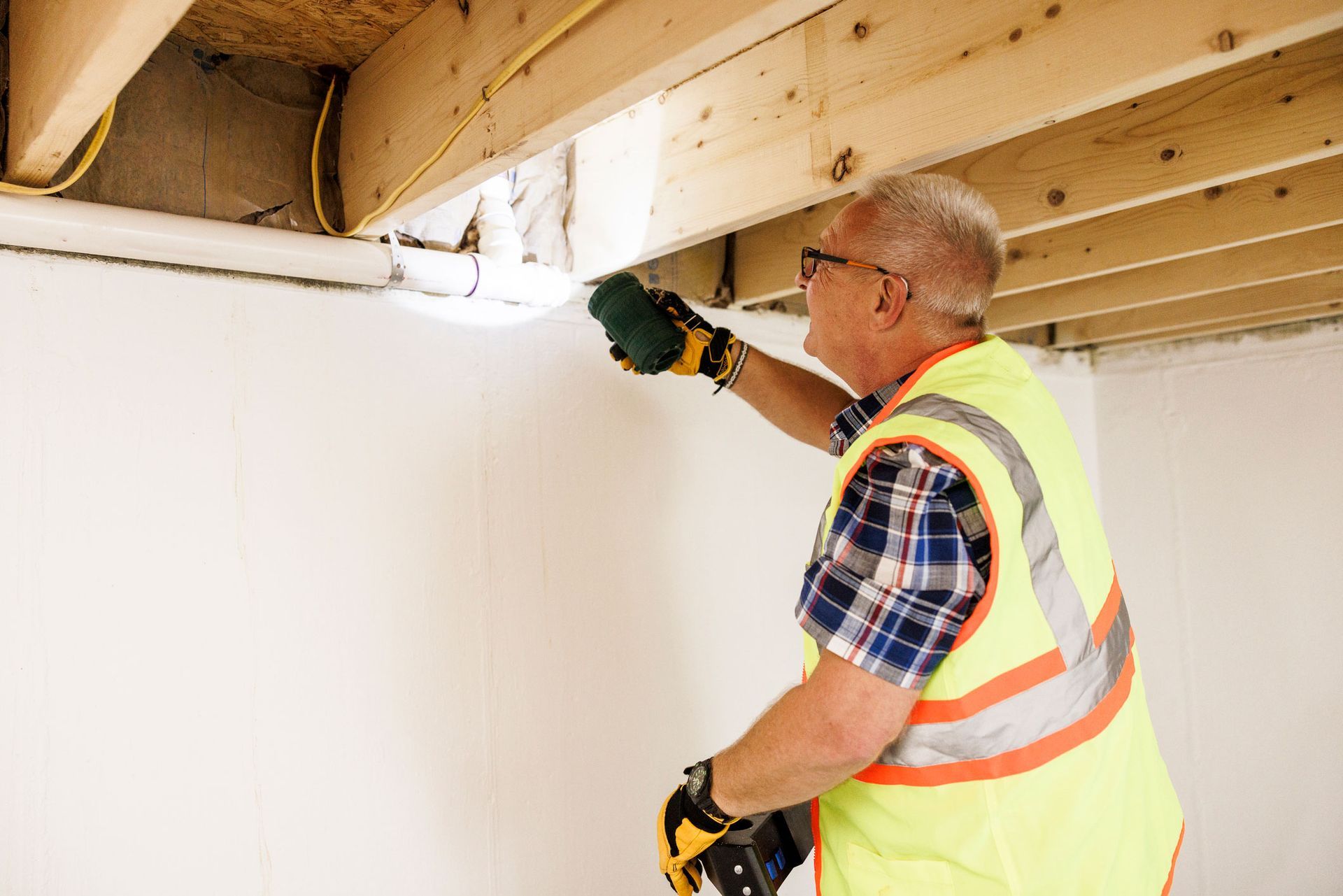 A man in a yellow vest is working on a wall in a basement.
