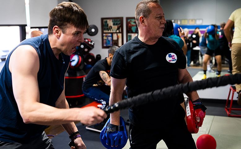 Men in a gym, exercising. One man is yelling while holding a black rope, another has boxing gloves.