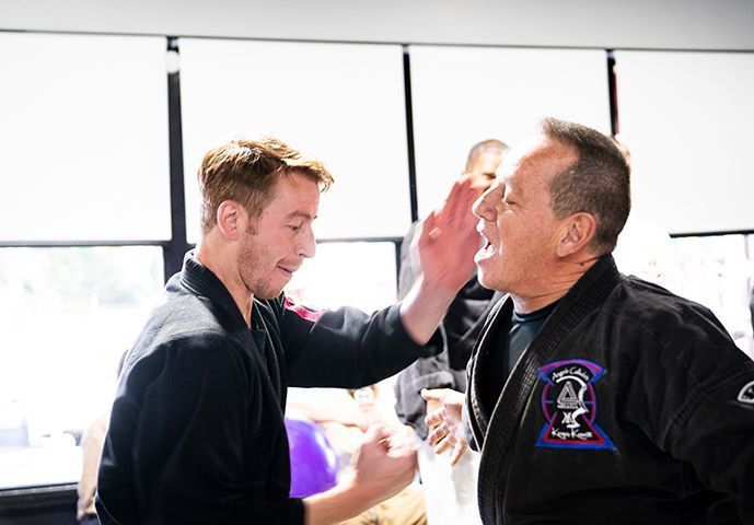 Man in black uniform slaps another man in training. Indoors, other people watch.