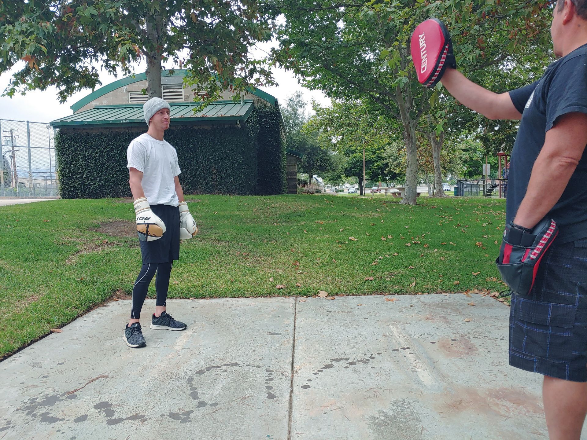 Man in black leggings and gloves, practicing boxing with a trainer outside, holding mitts.