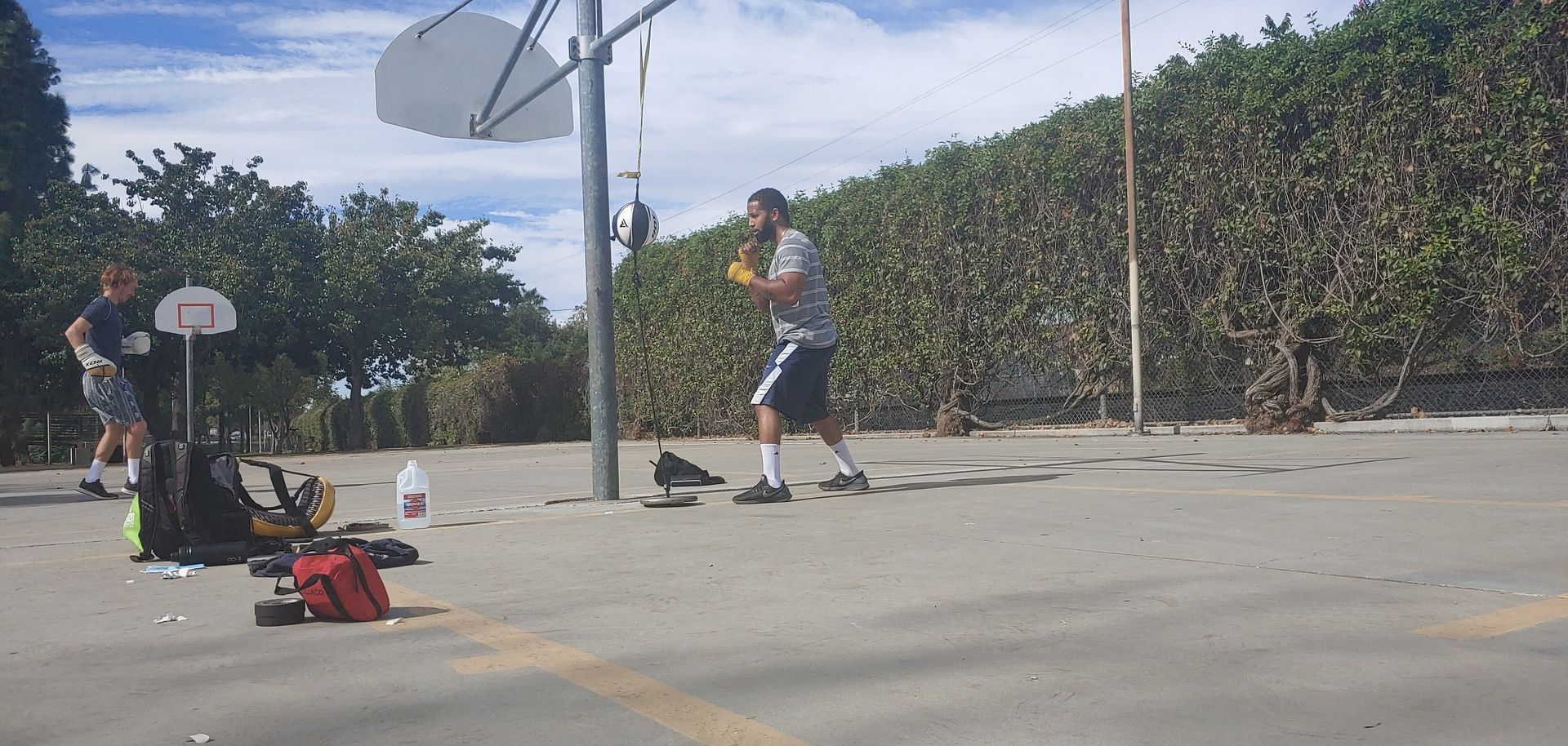 Man dribbling basketball on outdoor court, another person standing by a hoop, equipment on the ground, green hedge in the background.