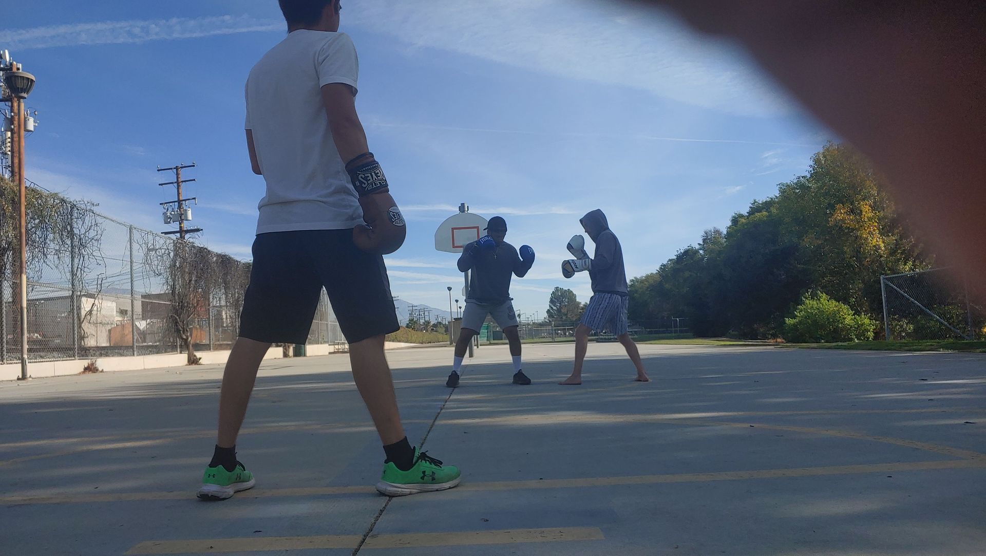 Three people boxing on a basketball court. One watches while two spar. Bright blue sky.