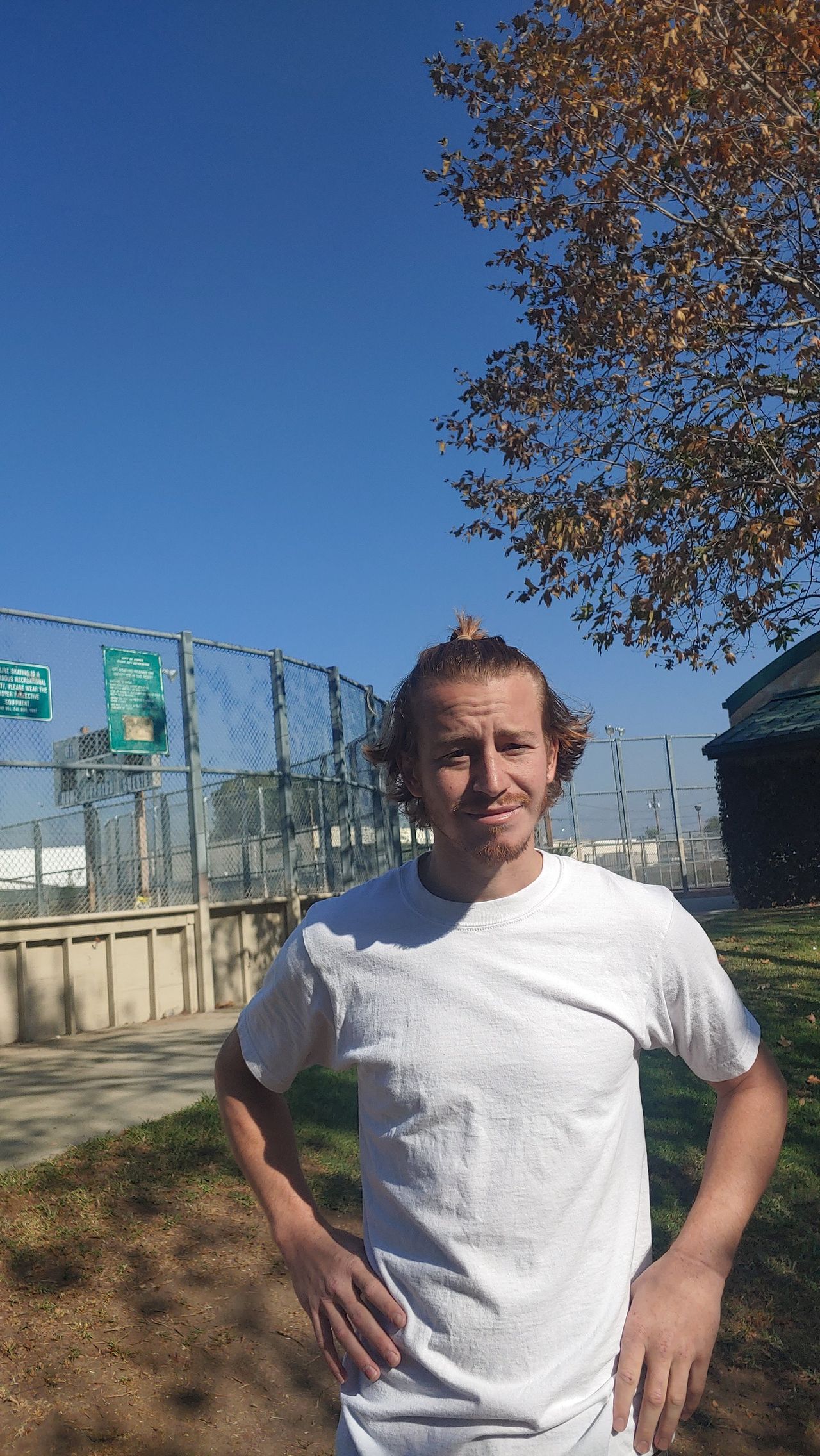 Man with curly hair and white shirt, hands on hips, standing outdoors near a fence and trees on a sunny day.