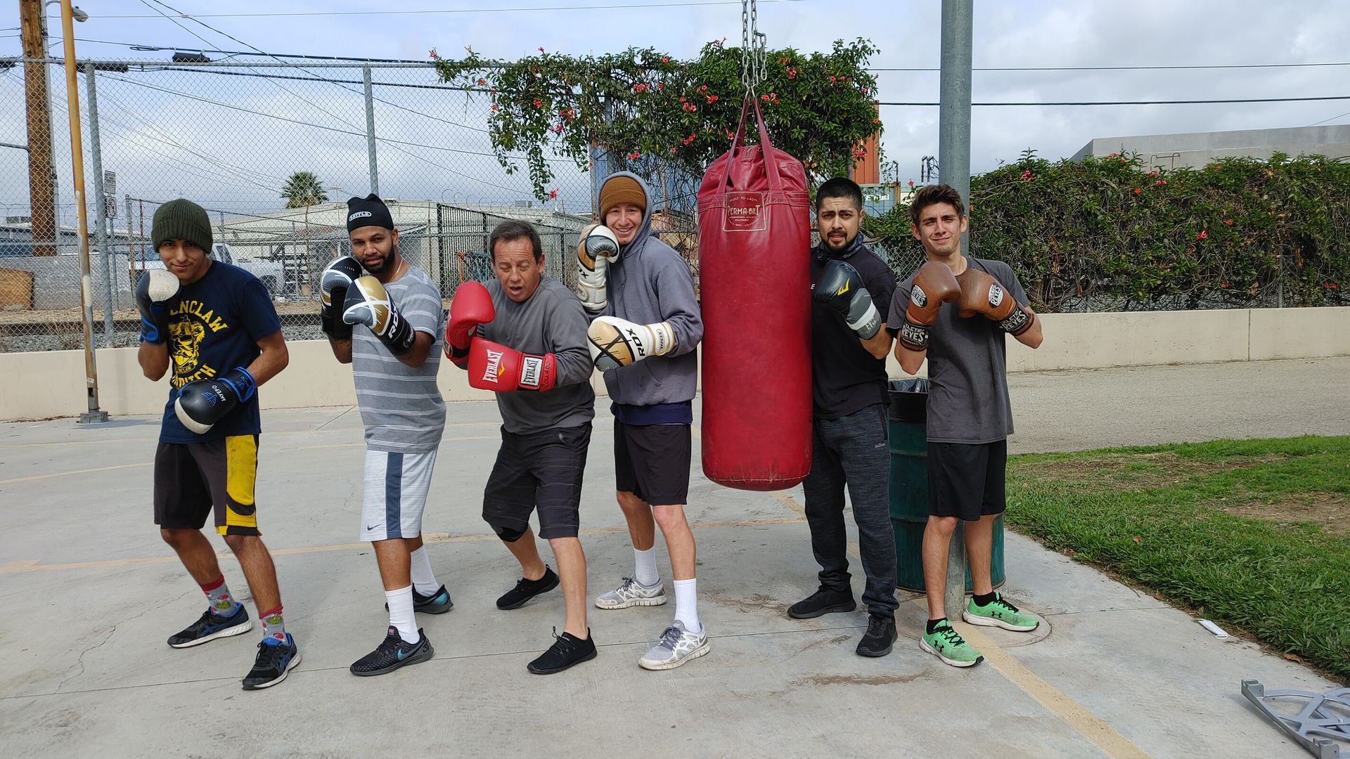 Six people in boxing gear pose by a red punching bag, outdoors. They wear gloves and athletic clothing.