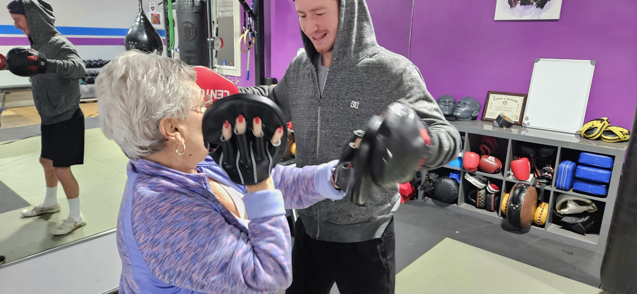An older person and a coach boxing in a gym. The coach holds mitts, while the other person punches. Purple wall.
