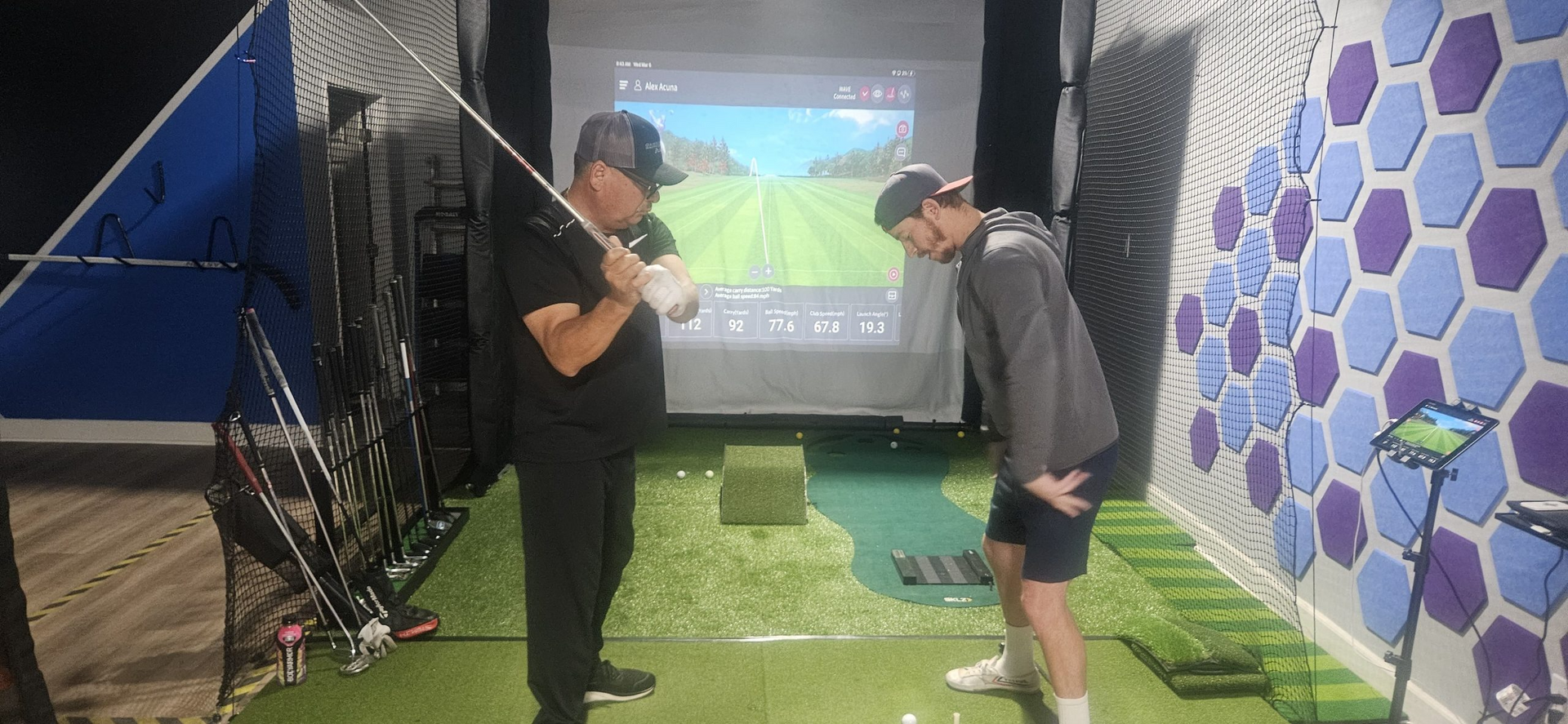 A man is teaching another man how to play golf in a golf simulator.