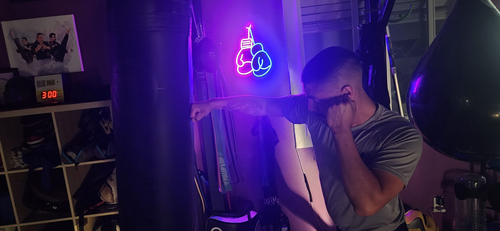 Man shadowboxing near a neon boxing glove sign.  He's wearing a gray t-shirt. The setting is dimly lit.