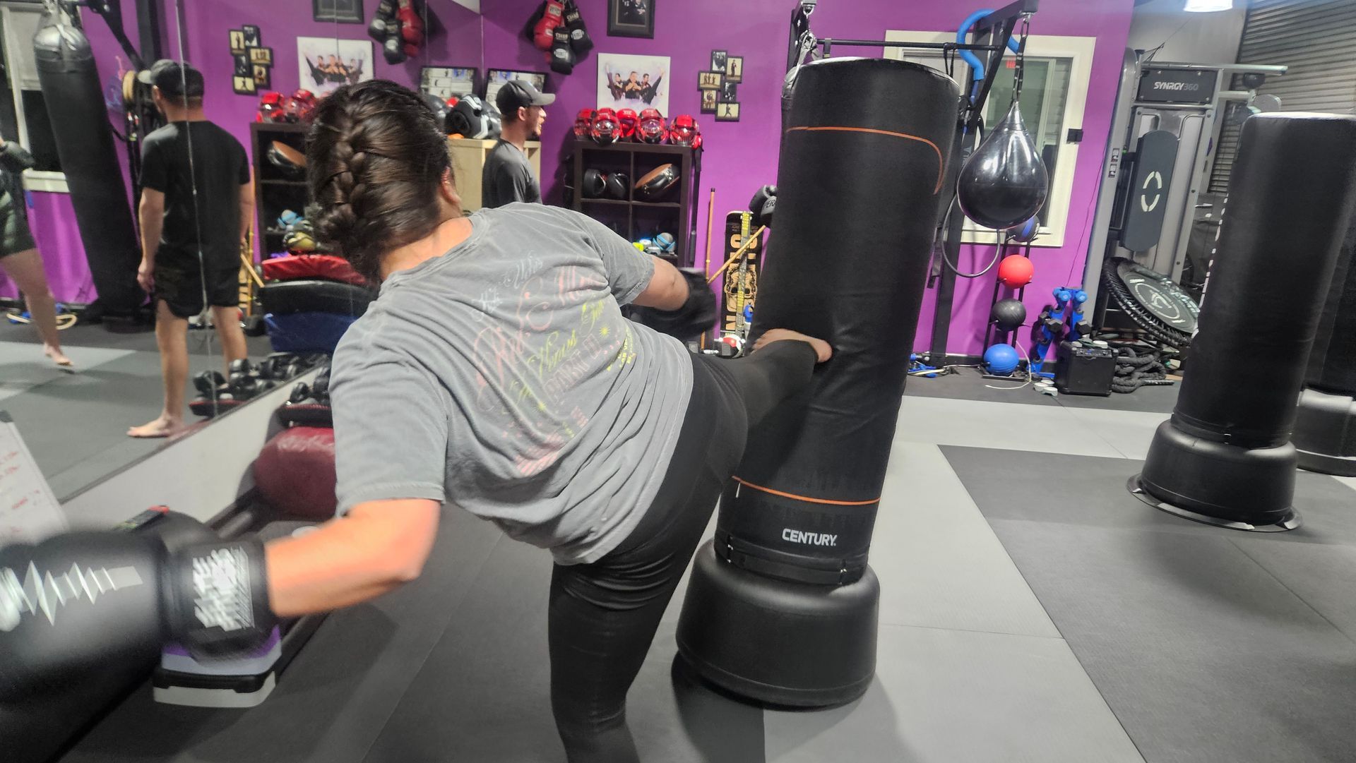 Woman practicing kickboxing on a gym mat, wearing gray and black gear, with punching bags in the background