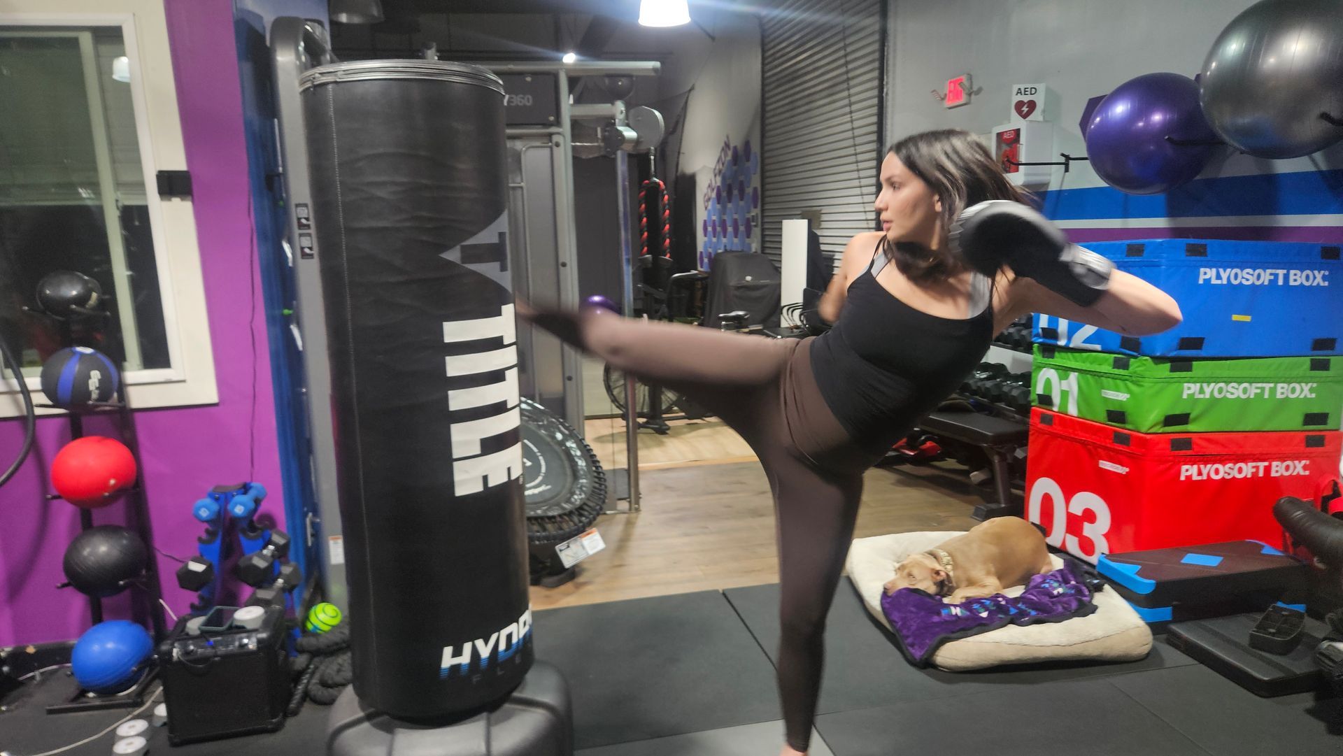 Woman kicking a tall black punching bag in a gym, with exercise equipment in the background.