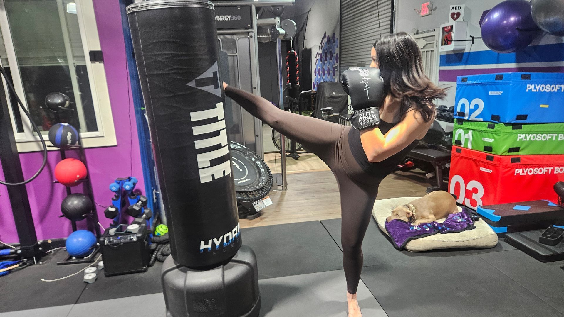 Woman in black workout clothes kicks a heavy bag in a gym with purple and red equipment.