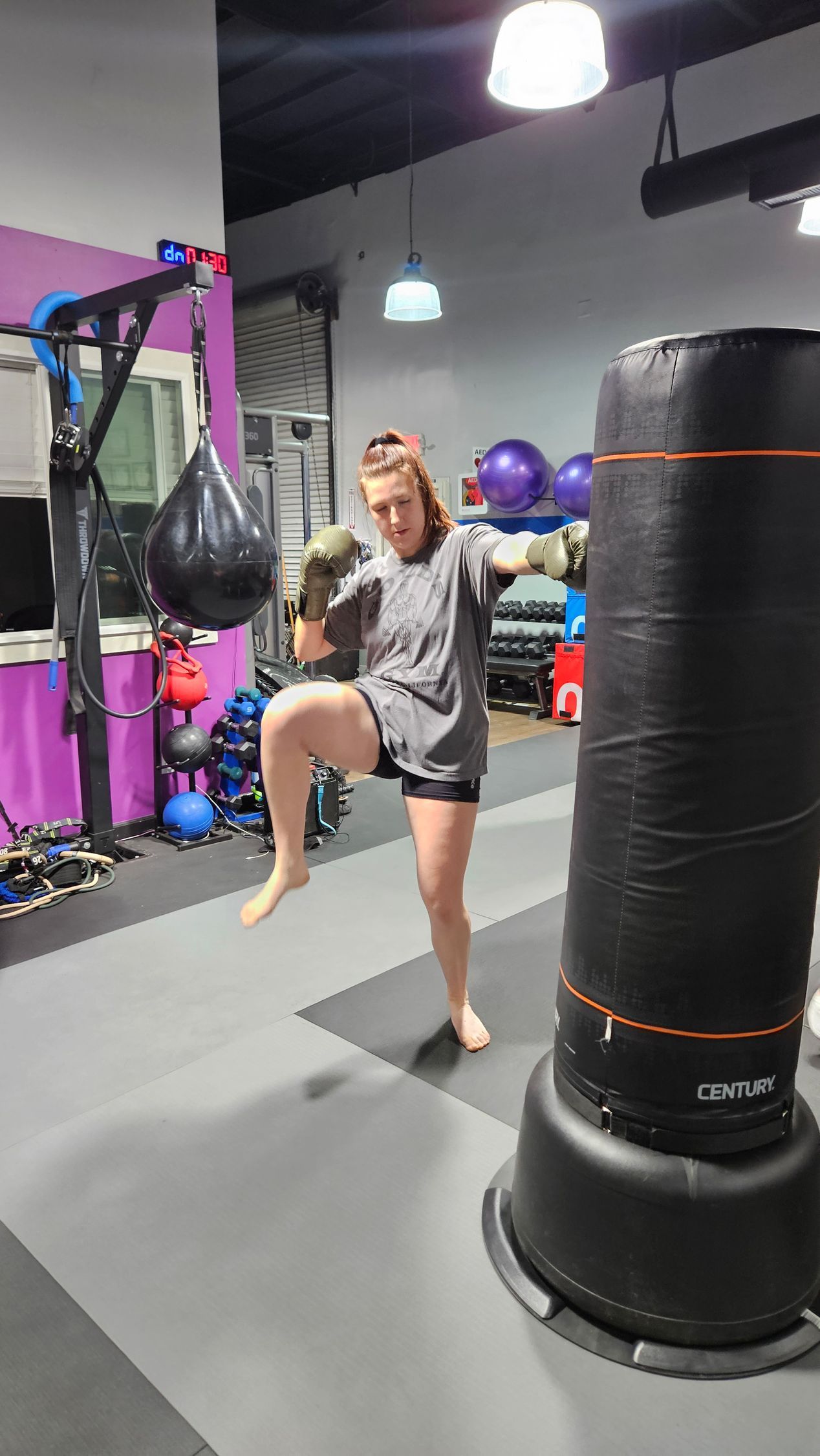 Person in a gym raises one knee beside a black punching bag, with colorful exercise equipment in the background
