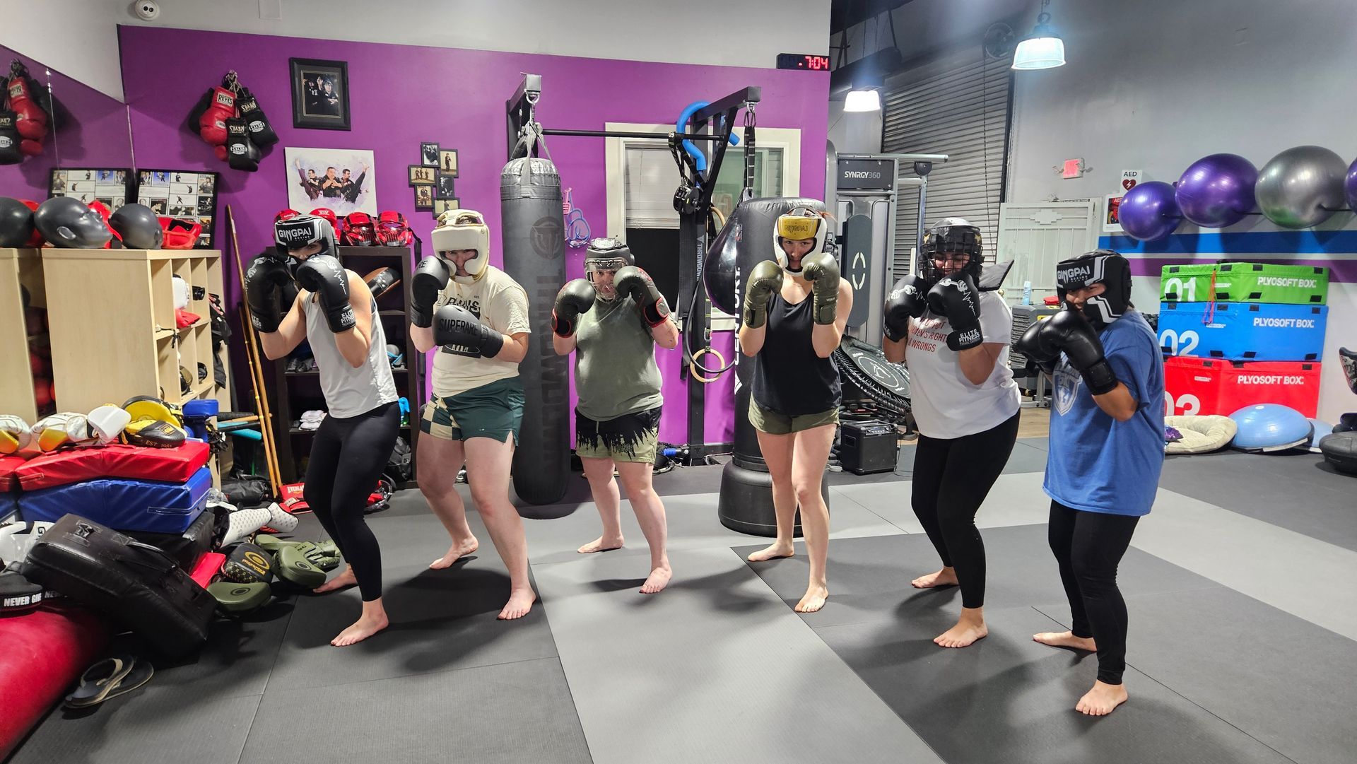 People posing with boxing gloves in a colorful gym, standing barefoot on padded flooring.