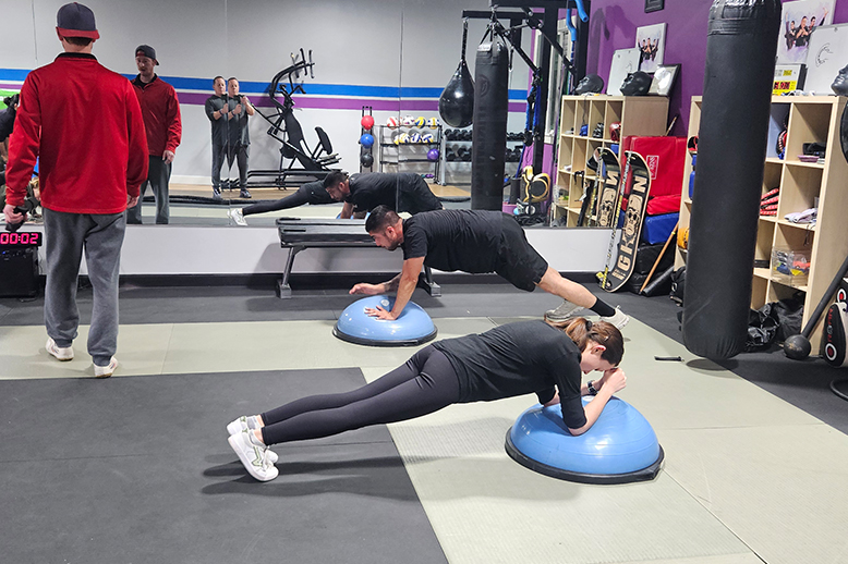 People in gym, doing planks on blue balance balls. Mirror reflection shows similar activity.