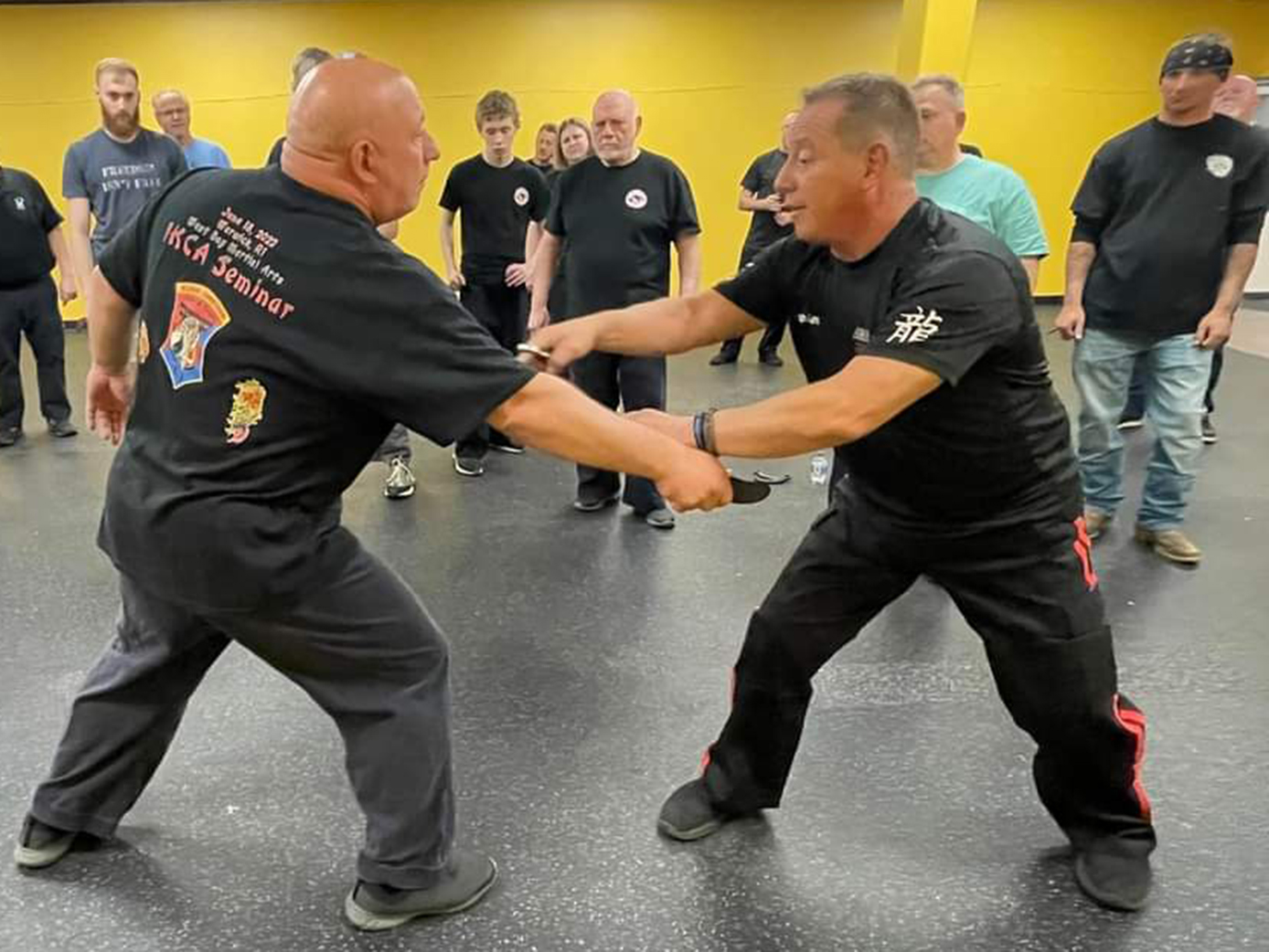 Two men in black shirts practicing knife defense, indoors, other people watching.