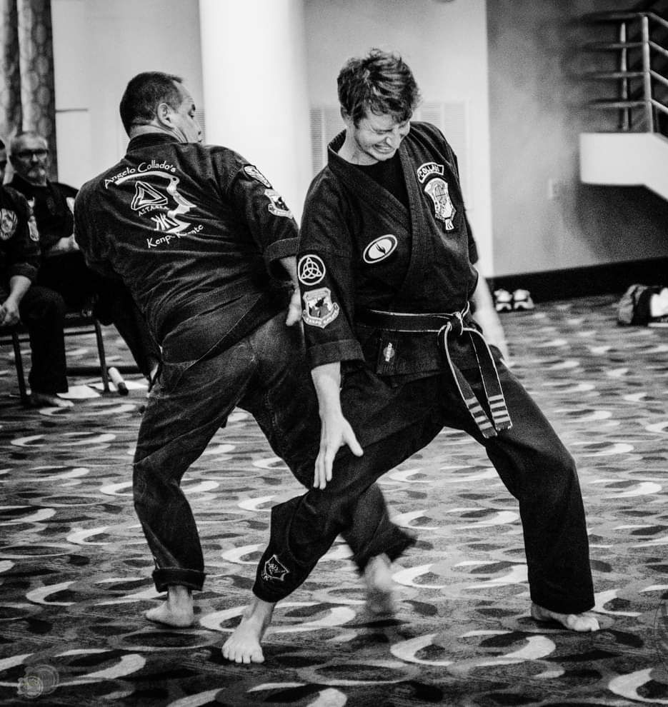 Two people in martial arts uniforms practice a block/kick in a carpeted room.