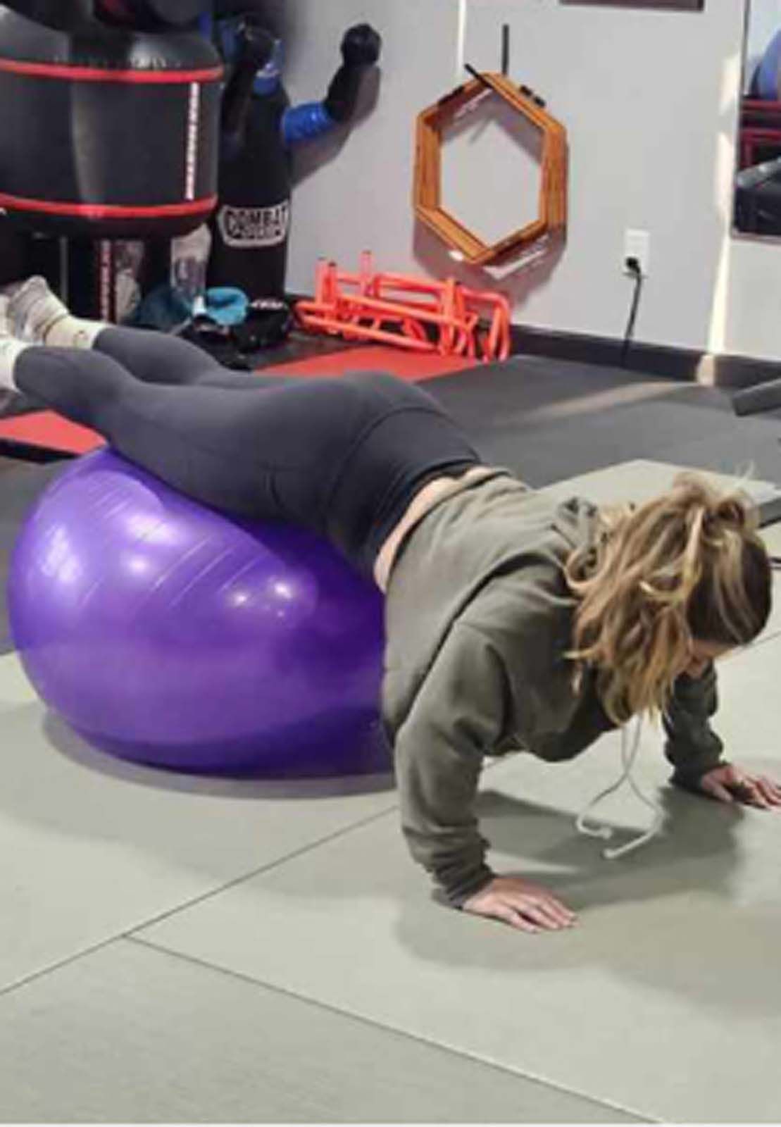 Woman performing push-ups on a purple exercise ball in a gym. She's wearing a green hoodie and black leggings.