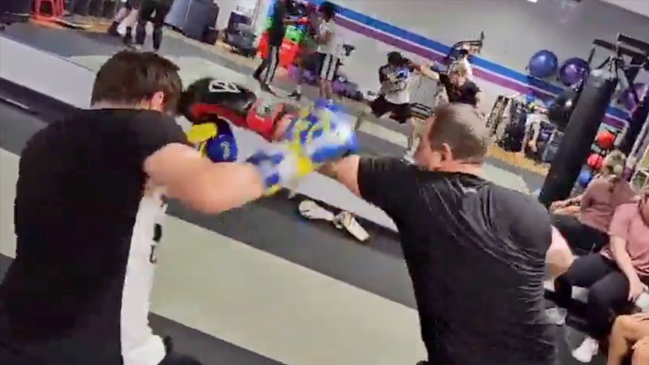 Two people sparring with gloves in a boxing gym, with onlookers and training equipment in the background