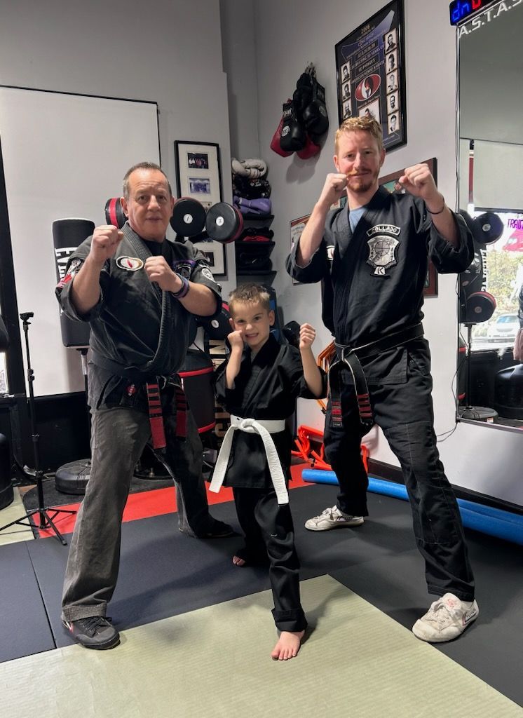 Three people in martial arts attire posing: two adults and a child, indoors, fists up.