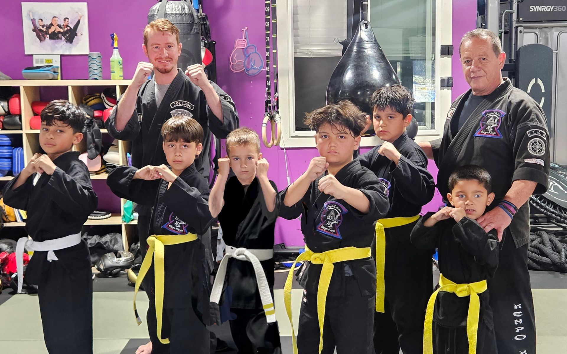 Martial arts class: Adults and children in black uniforms with yellow belts, fists up, indoors.