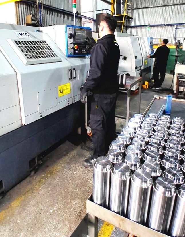 Un obrero maneja una máquina grande en una fábrica, con filas de piezas metálicas apiladas delante.