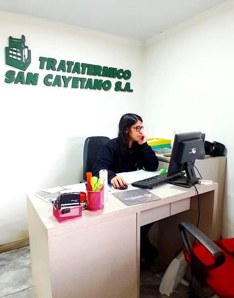 Mujer trabajando en un escritorio en una clínica con un letrero de “Tratamiento San Cayetano SL” en la pared.