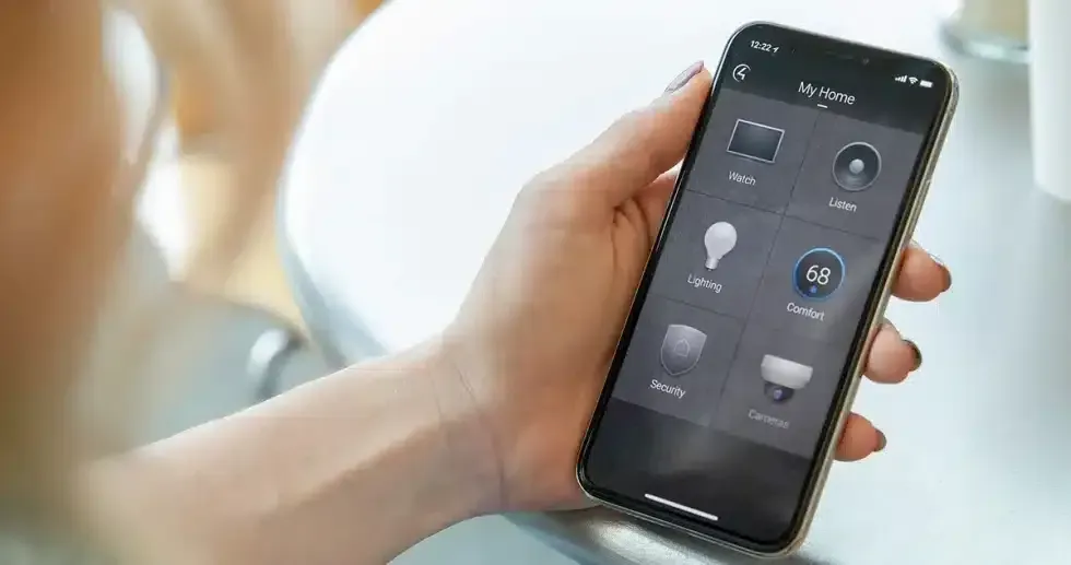 Person holding a smartphone displaying a smart home control app with various device icons.