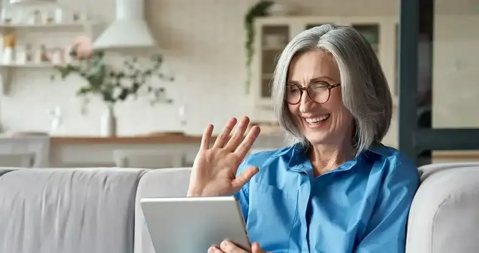 Woman with gray hair, wearing glasses, waving while using a tablet on a couch indoors.