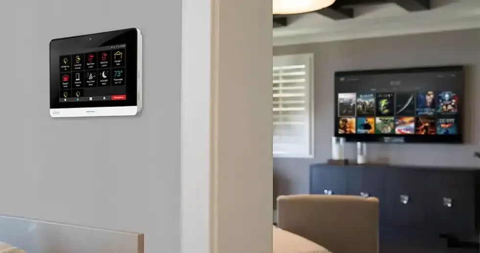 Smart home control panel on a gray wall, visible in a living room with a TV and furniture.