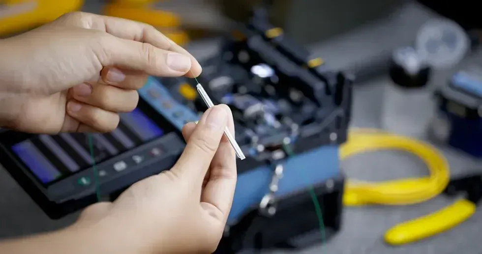 Hands holding fiber optic cable, preparing for a fusion splice with tools in the background.