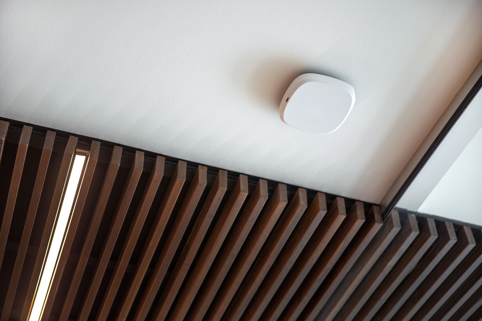 White ceiling with a smoke detector, above brown wooden slats with a vertical light.