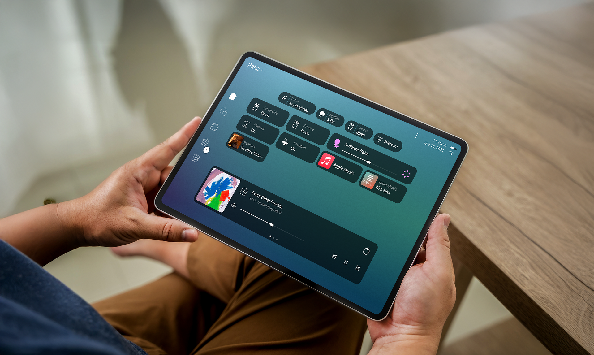 Hands holding a tablet displaying a home control interface with a wooden table in the foreground.
