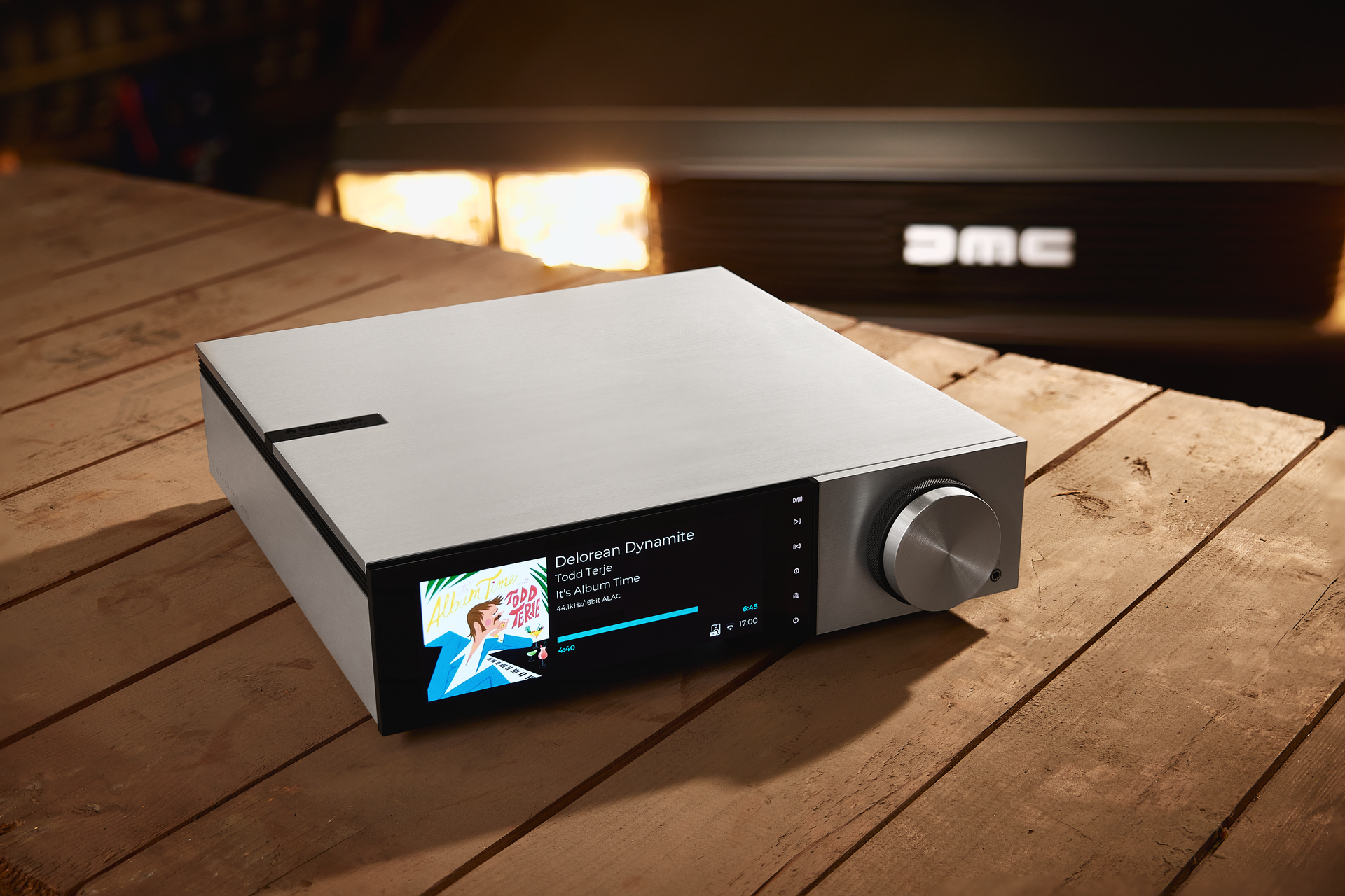 Silver audio receiver on a wooden surface with a car in the background displaying song information on its screen.