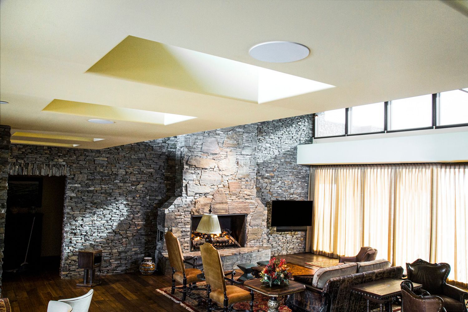 Living room with stone fireplace, skylights, and seating around a table, with curtained windows.
