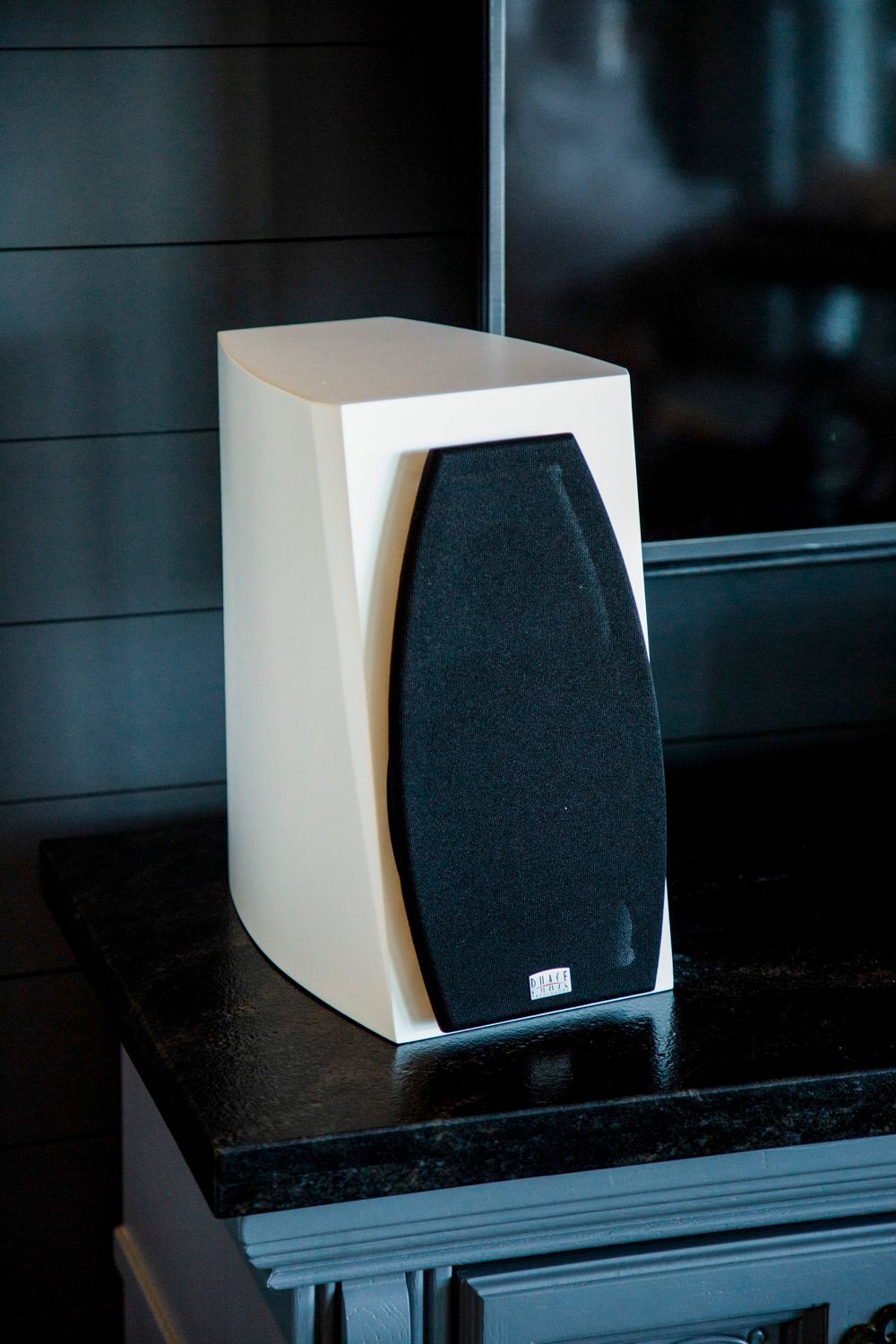 White and black speaker on a black surface against a dark gray wall.