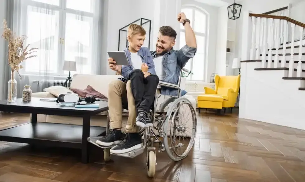 Man in wheelchair celebrates with boy looking at tablet in bright living room.