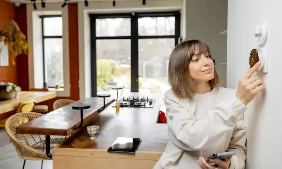 Woman adjusting a smart thermostat on a white wall in a modern kitchen. She holds a phone.