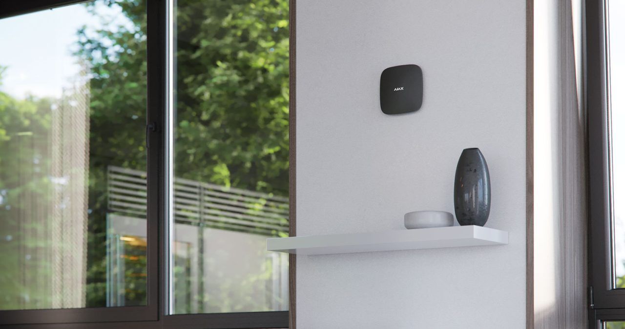 White wall with shelf holding vase and a smart device, window view of trees.