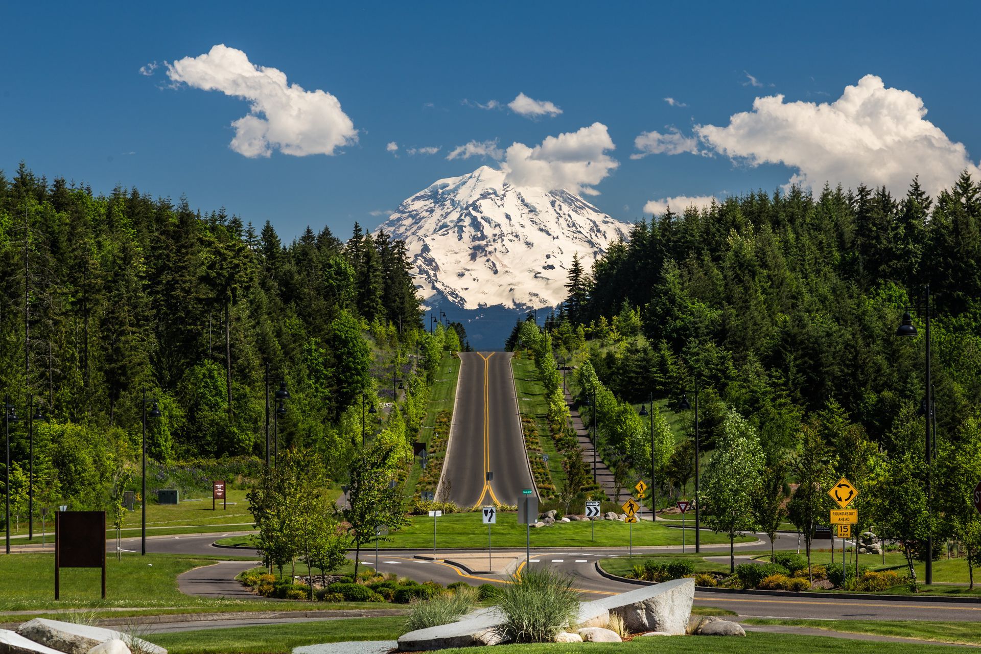 Road through a green forest leading toward a snow-capped mountain under a blue sky with clouds.