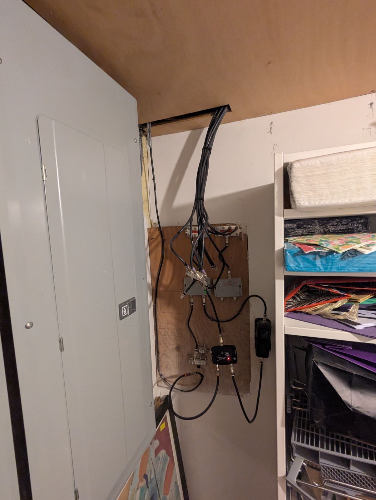 Electrical panel and wiring mounted on a wooden board, near a closet with shelves and artwork.