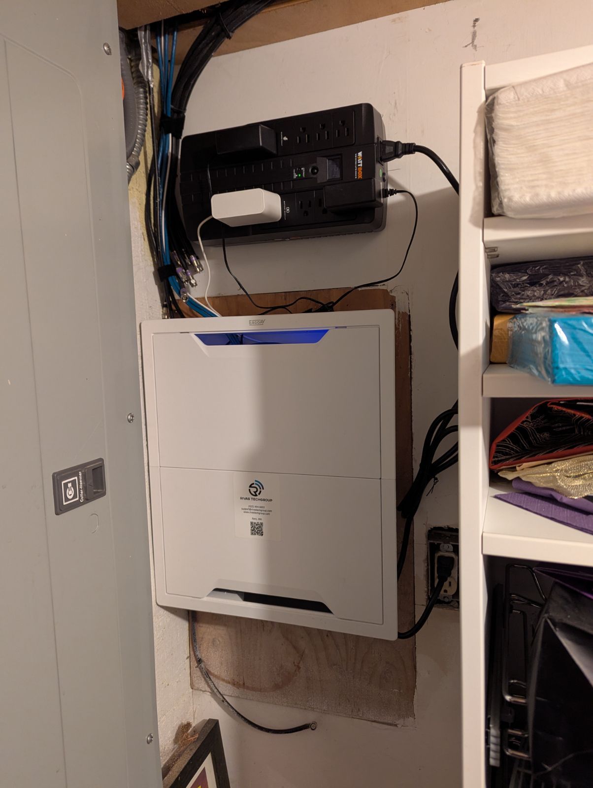 White box with blue light and black power strip mounted on a wall with wires and shelving.