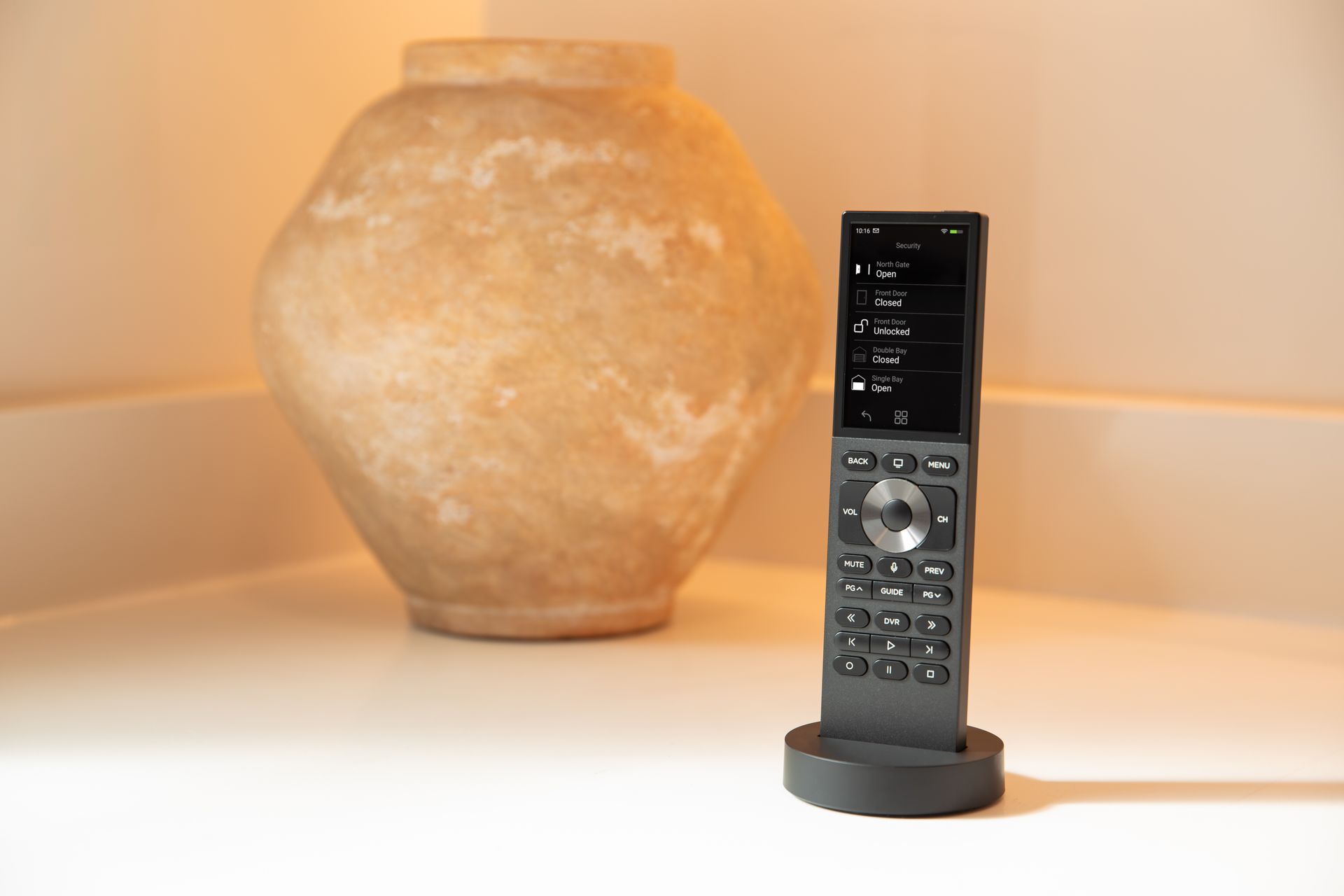Remote control in a charging base on a white surface, with a blurred beige vase in the background.