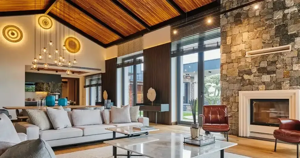 Living room with fireplace, sofa, and large windows. Wooden ceiling, stone wall, and modern lighting.