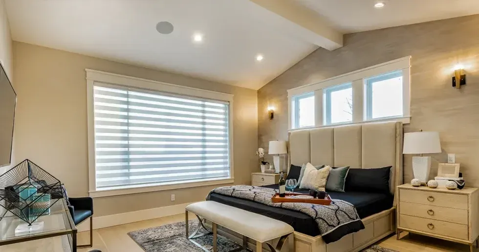 Bedroom with king-sized bed, large window with blinds, beige walls, and light-colored furniture.