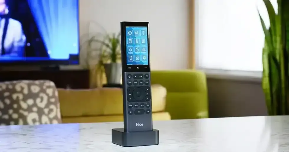Black smart remote with touchscreen in a charging dock on a table. In the background: TV and green armchair.
