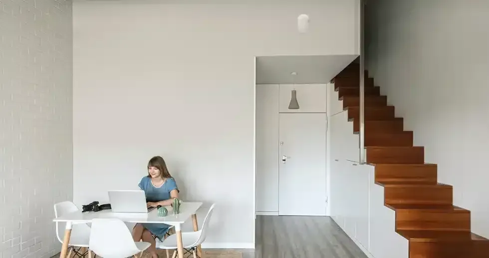 Woman working on laptop at table, light-filled room with stairs.