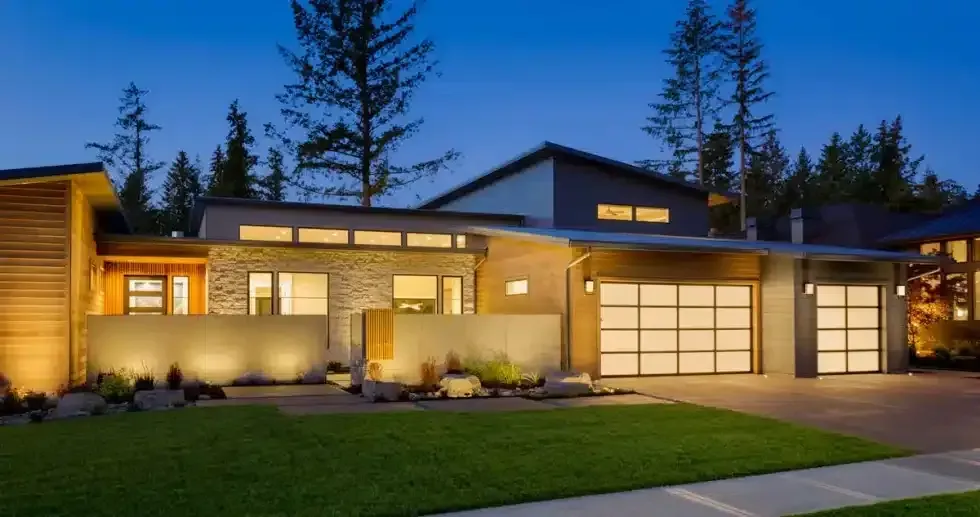 Modern home exterior at dusk, featuring a stone wall, garage doors, and green lawn.