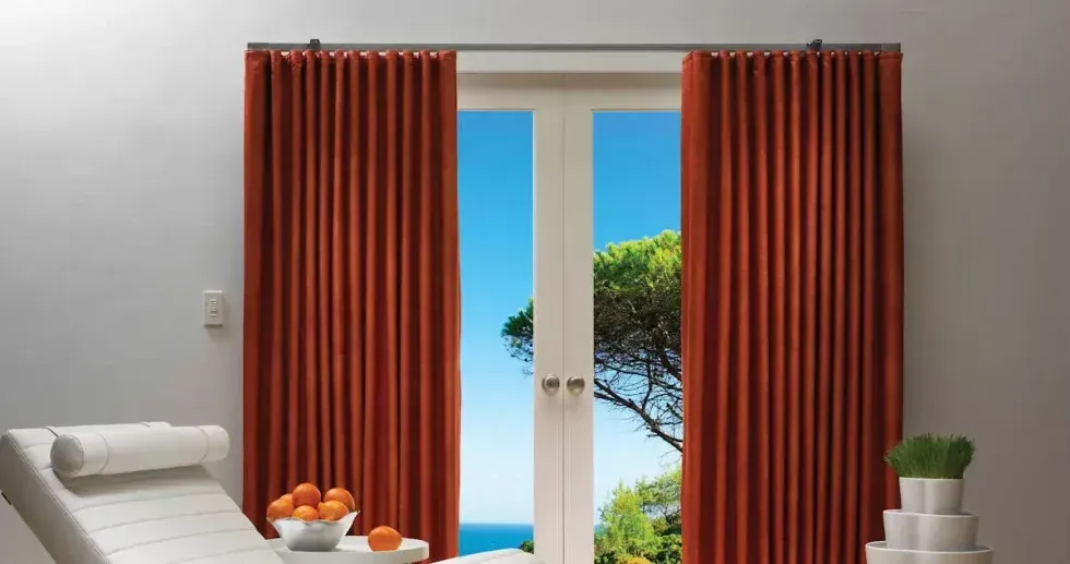 Orange pleated curtains framing a view of blue sky, ocean, and a tree, with a white chair and fruit in foreground.